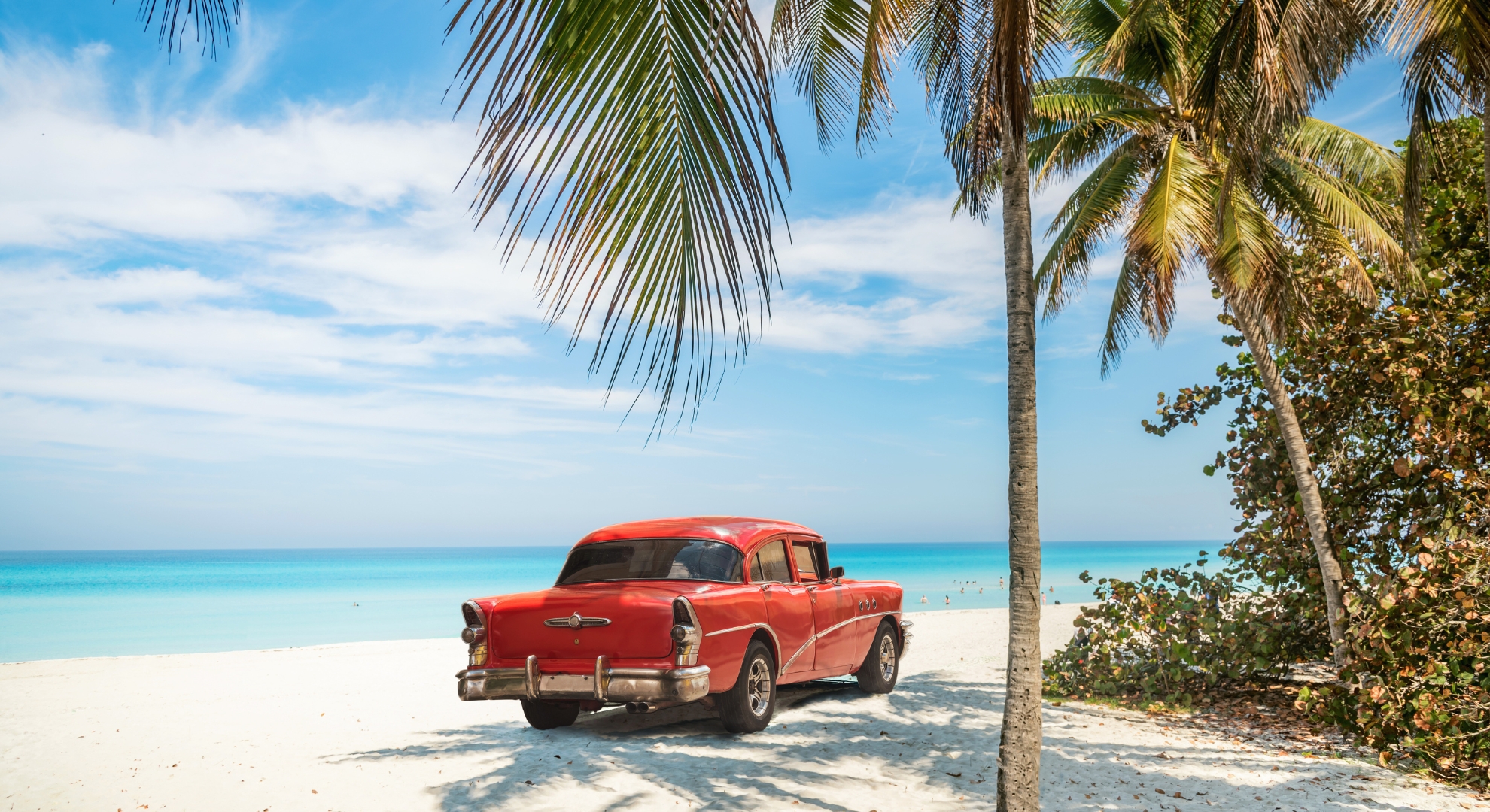 Classic car on a Cuban beach, surrounded by golden sand and turquoise water under a bright sky.