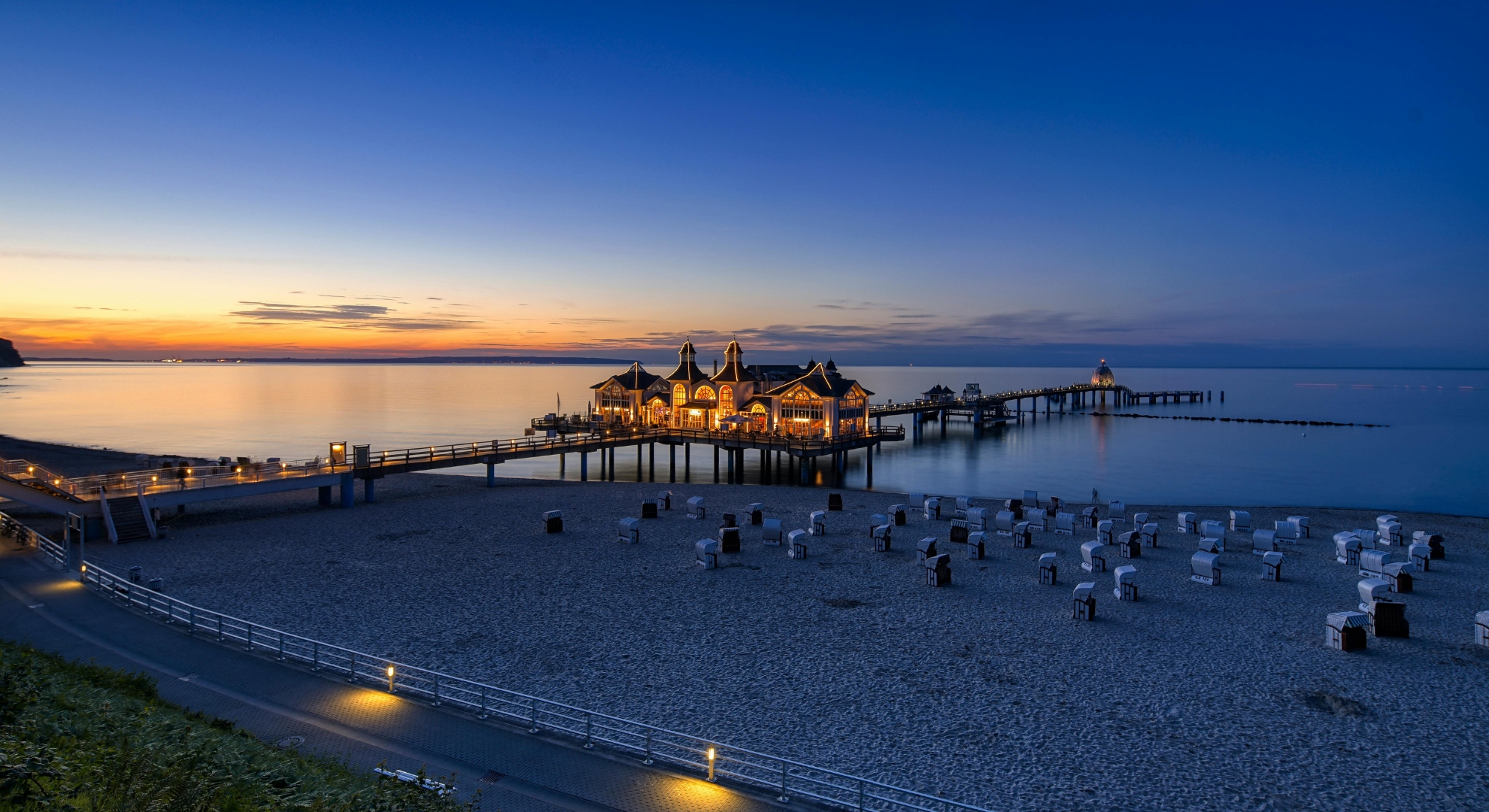 A sunset view of a German pier with chairs and a house, casting warm colors over the water.