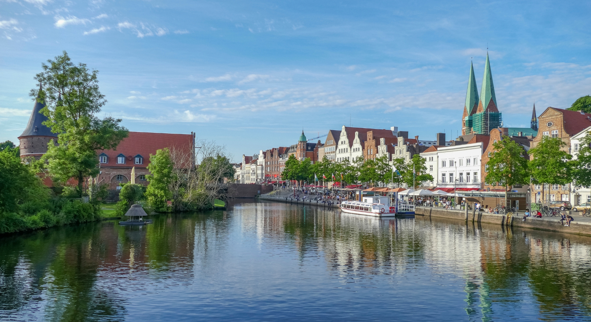 A river flows through a German city, surrounded by modern buildings and lush trees under a clear blue sky.