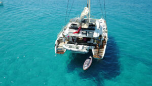 A catamaran, S/Y KASKAZI FOUR, sails gracefully in clear blue waters under a bright sky.